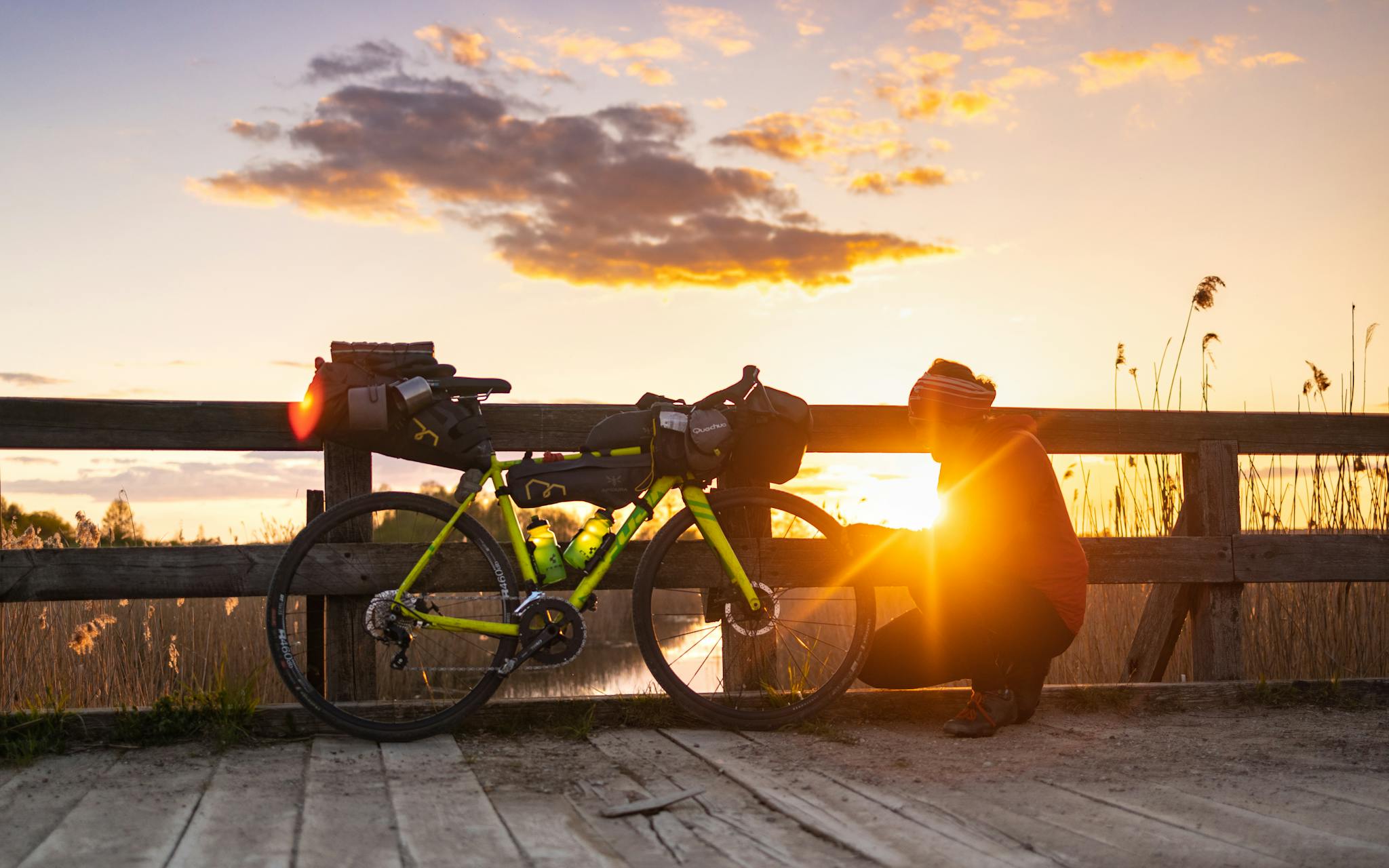 Cyclist resting beside a loaded bike during sunset on a scenic wooden bridge, capturing the essence of adventure.