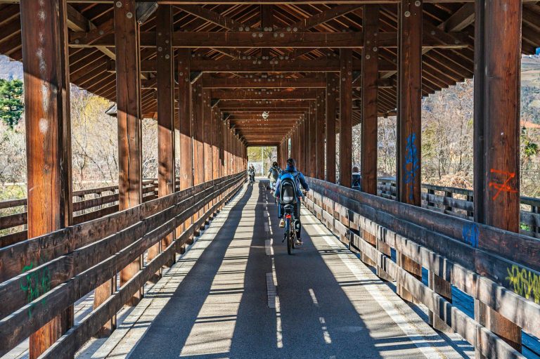 Cyclists ride through a wooden covered bridge on a sunny day showcasing perspective and adventure.