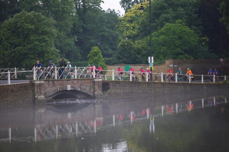 Cyclists riding across a bridge over a calm misty lake in a lush green park.